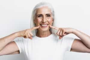 Femeie zâmbitoare, cu păr alb, arătând spre dinți, simbol al unui zâmbet sănătos după tratamentul cu implant dentar