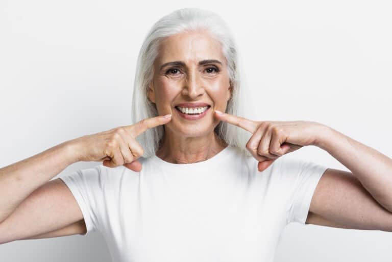 Femeie zâmbitoare, cu păr alb, arătând spre dinți, simbol al unui zâmbet sănătos după tratamentul cu implant dentar
